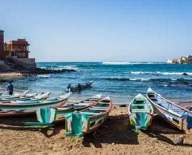 Colorful wooden fishing boats docked on a sandy beach in Senegal with ocean waves in the background.