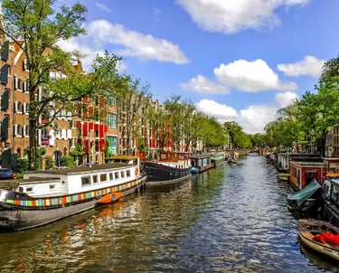 Traditional houseboats and brick buildings line a scenic Amsterdam canal under a blue sky.