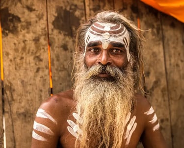 A Hindu Sadhu with traditional white Tilak face paint and a long beard poses against a wooden background.