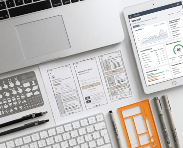 Mobile app wireframes and SEO dashboard displayed on a desk with a laptop, tablet, and design tools.