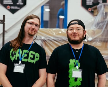 Two vendors with cool nerdy t-shirts stand smiling at their booth.
