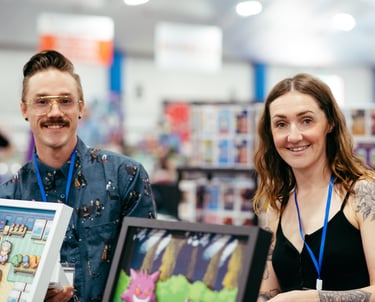 Two smiling vendors stand in front of the video game themed art they have for sale.