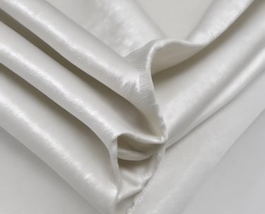 A close-up view of a carding machine with white wool fibers being processed. The fibers are aligned and spread out over a surface with fine metal teeth, with a wooden handle visible in the background.