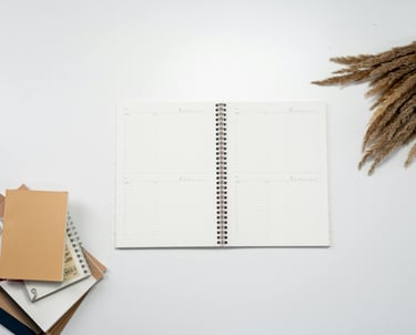 Flat lay of an open spiral notebook planner on a white desk with stacked journals and dried pampas grass.