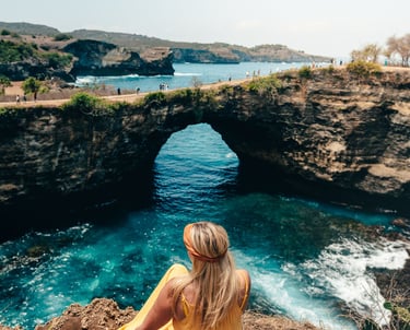 A blonde woman in a yellow dress sits overlooking Broken Beach in Nusa Penida, Bali.