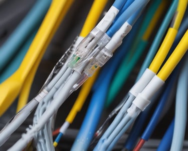 A person wearing protective gear and a welding helmet is engaged in welding activities, surrounded by various tools and equipment in a workshop environment. The bright light from the welding process illuminates the dark space, highlighting the focus and intensity of the work. The setting includes items such as gas tanks, a red cooler, and assorted tools on shelves.