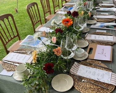 Long outdoor banquet table set with green linen, woven placemats, and a colorful floral centerpiece.
