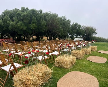 Rustic wedding ceremony setup with bamboo chairs, straw bales with carnations, and woven jute rugs on the grass.