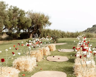 Rustic outdoor ceremony aisle with straw bales, red and white flowers, and woven jute rugs on a grassy lawn.