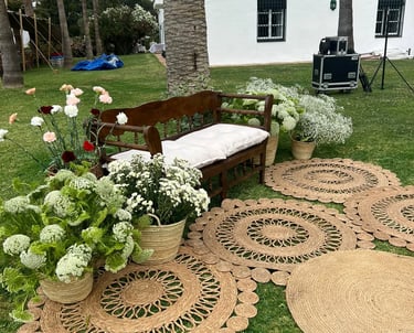 Rustic wooden bench with white cushion, surrounded by flowers and round woven jute rugs on a grassy lawn.