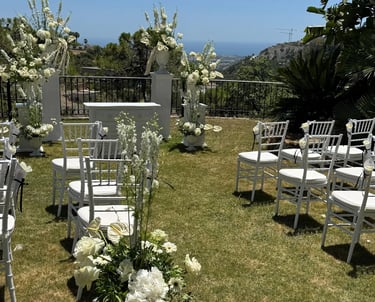 Outdoor wedding ceremony setup with white chairs, lush floral arrangements in white urns, and a scenic ocean view.