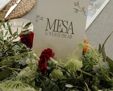 Elegant wedding table centerpiece with green foliage, red carnations, and a card reading 'MESA LOVE IS IN THE AIR'