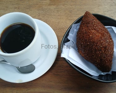 Uma xícara de café fumegante sobre um pires branco, ao lado num pratinho, um kibe dourado e crocante