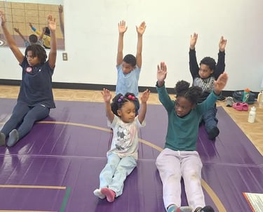 Young children sitting on a purple gym mat stretching their arms up during a group fitness class.