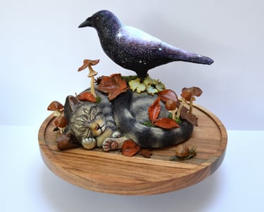 Handcrafted sculpture of a celestial galaxy crow perched on a sleeping tabby cat figurine with autumn leaves and mushrooms.