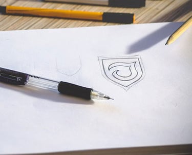 A drawing of a shield on paper, surrounded by pencils and a pen on a wooden surface.