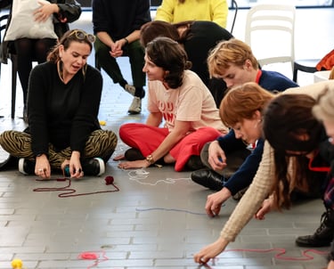 Eight adults sitting on the ground making patterns with colourful yarn.