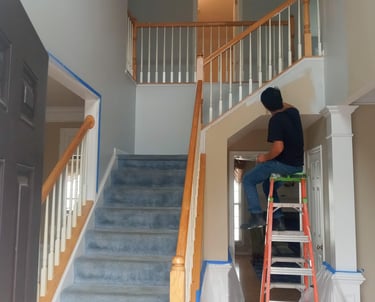 A person working on home renovation, standing on a ladder in a room with white walls and a staircase