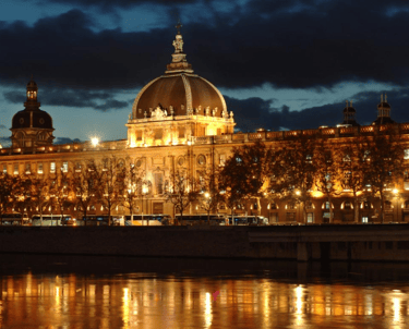 Vue nocturne de l'Hôtel-Dieu de Lyon illuminé, avec ses lumières dorées se reflétant dans le Rhône.