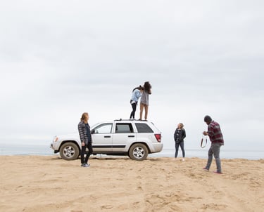 A group of friend in a beach. A car is parked on the beach and the group is posing for a picture