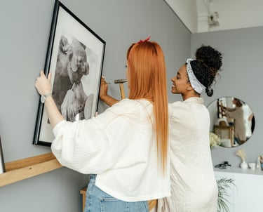 Two women installing framed refugee artwork in a gallery space.