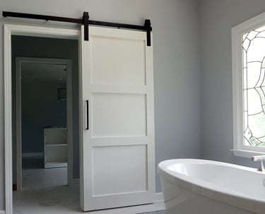 White 3-panel sliding barn door with black hardware installed in a modern gray bathroom.