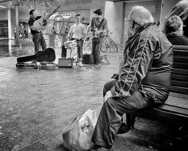 Older man sitting on mall bench listening to busking rock and roll group