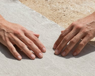 Close-up detail of hands delicately touching a swatch of raw greige linen fabric, sand-colored stone in the background, bright natural lighting, Spanish-speaking context.