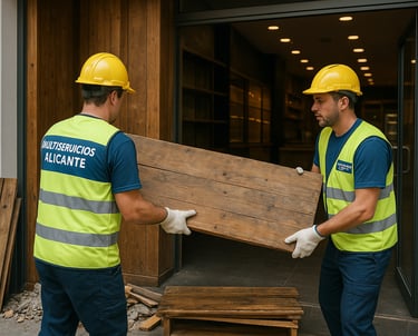 "Dos trabajadores de Multiservicios Alicante retirando una tabla de madera del interior de un local