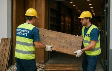 Dos trabajadores portando una madrea de un local y equipados con el traje de seguridad de la empresa