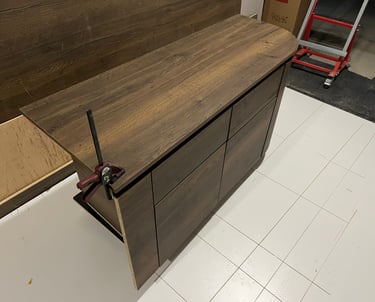 Dark oak wood laminate dresser being assembled with a bar clamp on a white tiled floor.