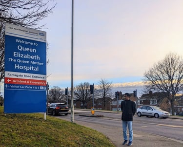 A man standing at a distance looking at the signpost reading 'Welcome to Queen Elizabeth The Queen Mother Hospital'