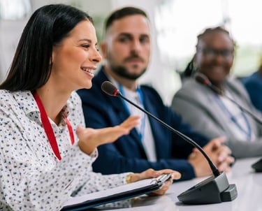 A smiling female speaker using a gooseneck conference microphone during a formal panel discussion, seated alongside two other