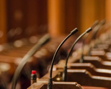 A row of gooseneck microphones installed on individual wooden podiums inside a formal conference or parliamentary setting, wi