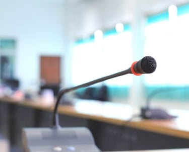 A close-up of a gooseneck conference microphone with a red ring around the mic head, placed on a wooden table in a brightly l