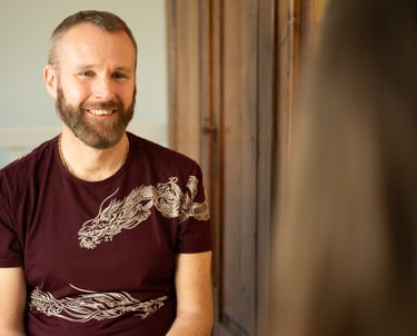A man wearing a dark red t shirt with dragon imagery is sitting coaching a client