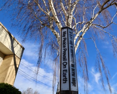 A White and black Peace pole with the word "May peace prevail on earth under a bright blue sky and the birch tree.