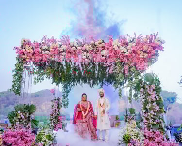 Indian bride and groom at an outdoor wedding ceremony under a floral mandap with colorful smoke.