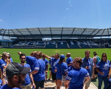 Crowd of fans in blue shirts socializing on the field at Sporting Kansas City stadium under a bright sky.