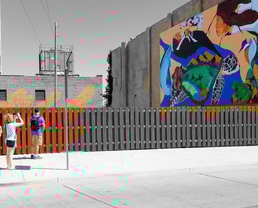 People photographing a colorful mural on a city building, with the surrounding street and structures in black and white.