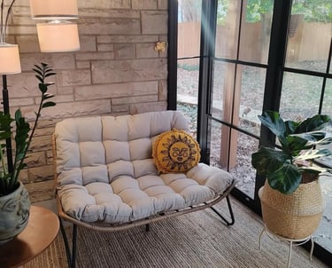 Cozy reading nook with a tufted papasan loveseat, sun pillow, and potted plants by a large window.