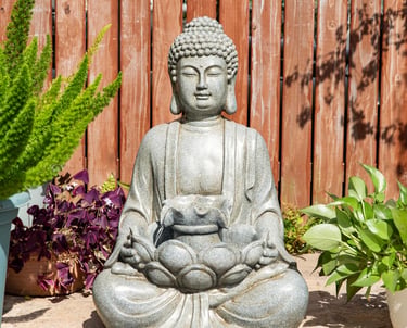 A stone Buddha garden statue sitting in a lotus pose near green plants and a wooden fence.