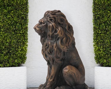 Majestic bronze-finished lion garden statue sitting between green hedges against a white wall.