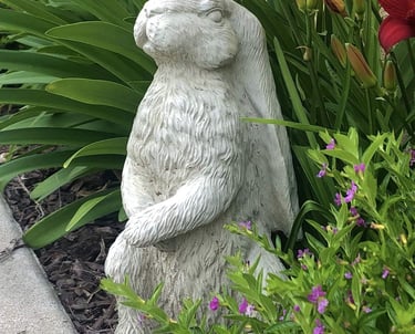 A decorative stone rabbit statue sitting in a garden among green leaves and purple flowers.