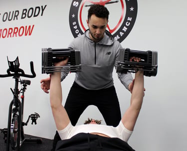 A personal trainer spots a man performing a chest press with adjustable dumbbells in a gym.