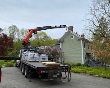 A knuckle boom crane truck delivering roofing materials to the roof of a green two-story residential house.