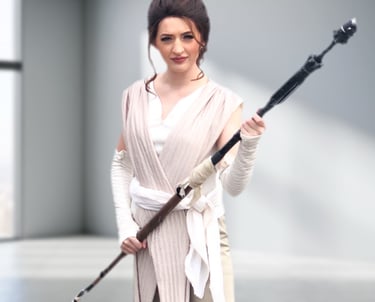 A woman in a realistic Rey Skywalker cosplay holding a staff in a minimalist studio.