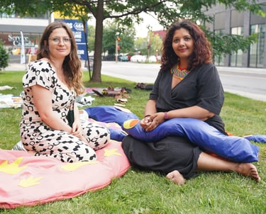 Two women pose for a portrait surrounded by soft body parts on the grass.