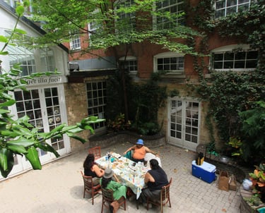 A group of four people sit outside on a hot sunny day sharing a meal in a plant-filled courtyard.