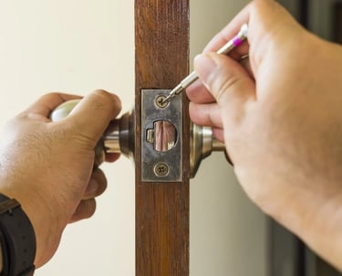 A person using a small screwdriver to repair a metal door latch on a wooden door frame.